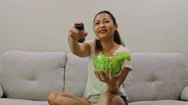 Neşeli Asyalı kadın, TV 'de eğlence şovu filmleri izlerken kanepede yeşil salata yiyor: Sağlık hizmetleri, yağ diyeti ve kilo verme kavramını azaltmak.