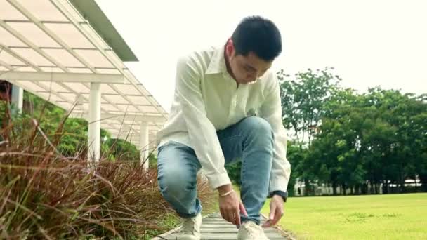 若いアジアの男ハンサムは 芝生で彼の緩い靴ひもを結ぶために歩いて停止し 良い気分でカメラを見る — ストック動画