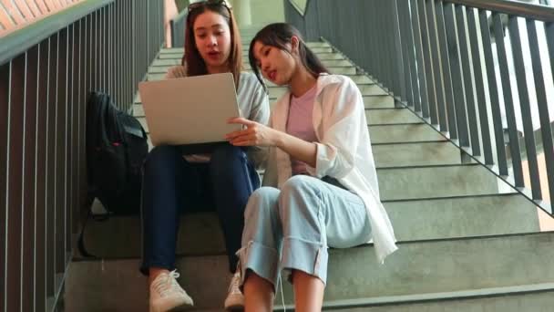 大学の2人のアジアの女性大学の学生は 興味を持って教室の階段に座ってラップトップ上の研究テーマを勉強しています — ストック動画