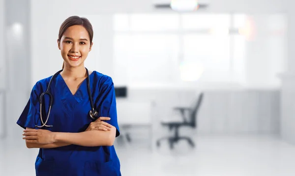 Portrait healthy asian female surgeon in blue dress with stethoscope standing crossed hands smiling friendly looking at camera standing smartly in abstract medical facility examination room.