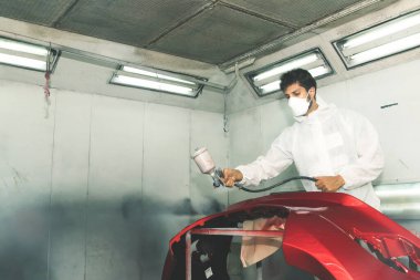 Otomotiv boya hizmetleri: Kırmızı sprey boya makinesi kullanan erkek boyacı, modern boya odasında tamir ve yenileme sürecinden geçen arabanın fiberglas ön tamponunda kırmızı boya spreyi.