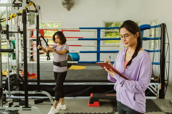 Happy portrait female personal trainer coach teacher working in gym holding clipboard recording workout giving advice as mentor correct and safe sports equipment to members private members.