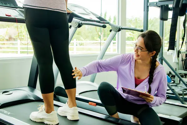Female coach trainer in fitness holding clipboard recording and giving advice and prevention of calf leg muscle injury during exercise, healthy young woman walking running on treadmill