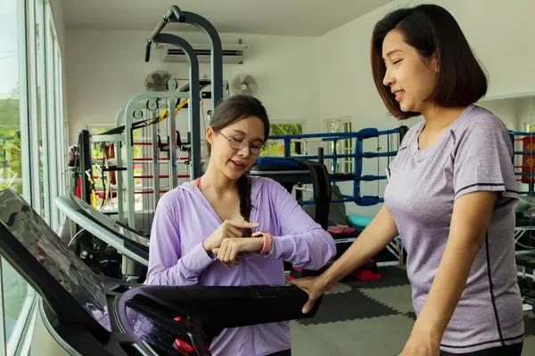 Female coach trainer gives exercise advice to young women who take care of their health by walking and running on an automatic treadmill and taking check timer to calculate their running properly.