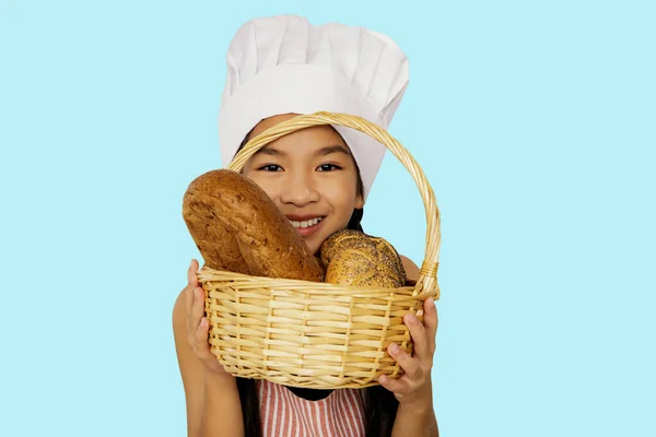 Happy asian young woman holding basket freshly delicious loaves bread wearing chef\'s hat on her head showing homemade bread made from wheat flour looking camera and smiling good mood blue background.