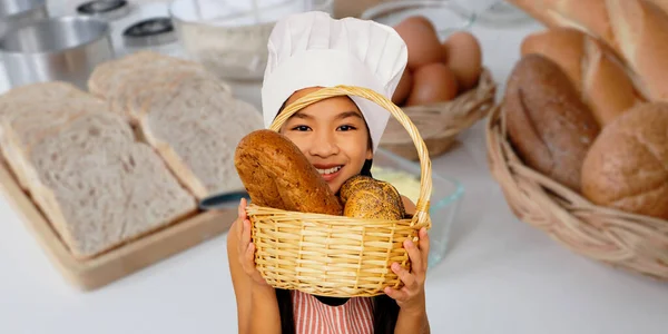 Happy portrait asian girl holding basket of freshly delicious loaves of bread wearing a chef\'s hat on her head showing homemade bread made from wheat flour and yeast.