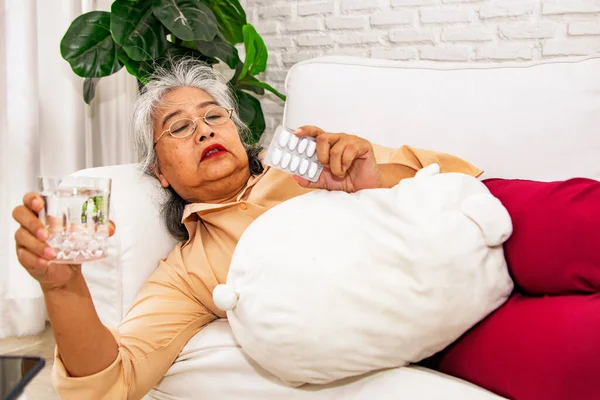 Asian elderly woman lying on the sofa, it\'s time to take medicine, holding glass of water and looking at medicine packets, wanting to take medicine every day to cure her own ailments.