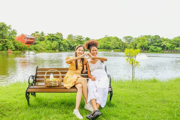 Portrait close friend asian woman having picnic in the park with healthy pregnant African American woman sitting on a wooden bench showing love, showing heart shape, looking cheerfully at the camera.