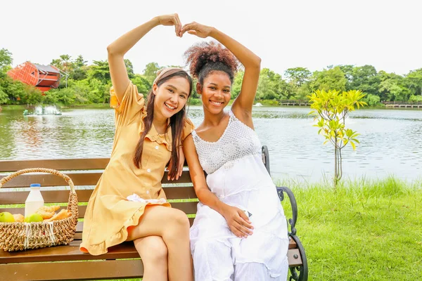 Portrait close friend asian woman having picnic in the park with healthy pregnant African American woman sitting on a wooden bench showing love, showing heart shape, looking cheerfully at the camera.