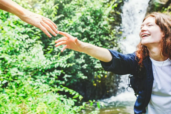 Male couple helps his girlfriend, reaching out to touch her walking across the rocks and streams waterfall in a tropical forest traveling with care expressing love and good wishes each other.