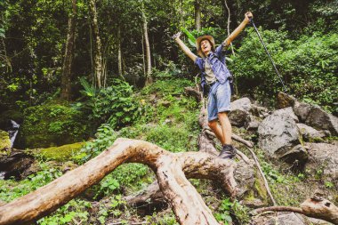 Neşeli genç adam, dağlarla, şelalelerle ve yemyeşil ormanlarla tropikal ormanlarda yürüyüş yapmayı seviyor. Kütüğün üzerinde dikilip güzelliği ve temiz havayı seyrediyor..