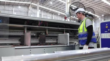 Erkek teknisyen bükülmüş karton kesme makinesi kullanıyor. Makine, lojistik karton üretim fabrikasındaki güvenlik ekipmanlarıyla güvenli bir şekilde çalışıyor..