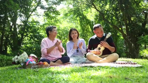 快乐的亚洲家庭外出野餐照顾他们的健康坐着弹奏四弦琴唱着赞美父母坐在一起支持家庭关系的女儿 — 图库视频影像