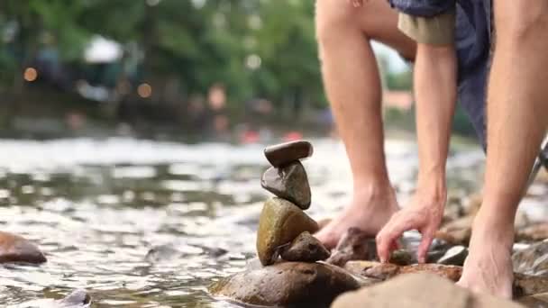 男子露营度假胜地的游客坐在河边 与一堆岩石一起玩乐 他把岩石筑成一座高塔 但不够稳固 不能倒在地上 — 图库视频影像