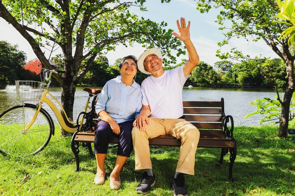 Senior asian retired couple healthy and cheerful travel health park parking bicycle sitting bench in an outdoor garden clear weather next shady pond looking at the beauty inside the amazing garden.