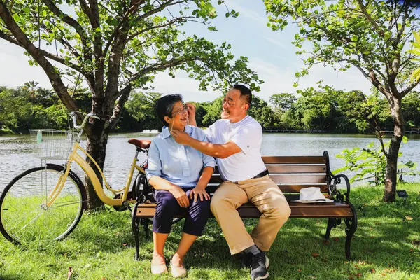 Senior asian retired couple healthy and cheerful travel health park parking bicycle sitting bench in an outdoor garden clear weather next shady pond looking at the beauty inside the amazing garden.