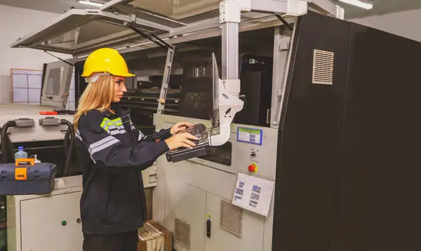Female worker wearing safety helmet operates keyboard controlling computerized automated Inkjet printing machine modern corrugated smart create program cardboard manufacturing logistics factory room.
