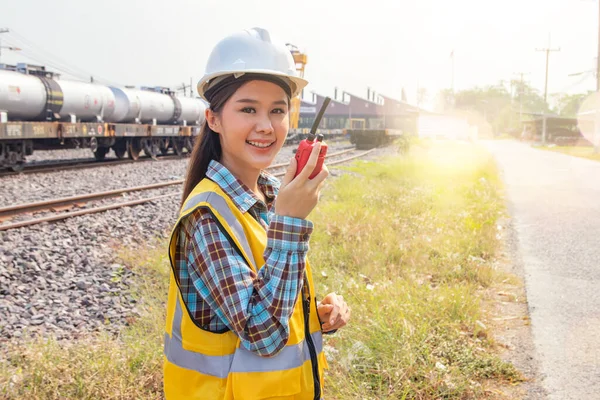 Beautiful asian woman engineer worker working in inspection field transporting diesel train crude oil transport rail into maintenance garage holding walkie talkie and helmet safety at train station.