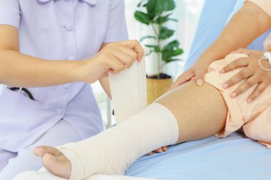 Kadın hemşirenin eli, ayak bileği burkulduğu için hastaneye kaldırılan yaşlı bir hastaya bakıyor bacağı ve ayak bileği ağrısını hafifletmek için bandaj kullanıyor:.