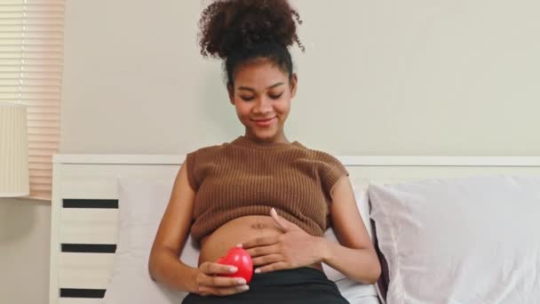幸せな妊娠したアフリカ系アメリカ人の母親は赤ちゃんとベッドに座って赤い心臓の健康な妊娠を示し カメラを見て笑顔 — ストック動画