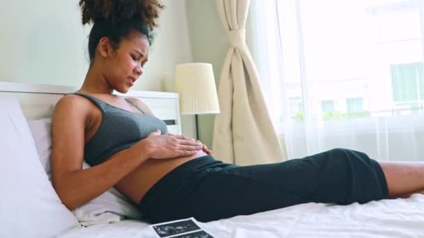妊娠中のアフリカ系アメリカ人の母親がベッドに座って赤ん坊を穏やかに脳卒中させ 赤ん坊が激しく労働苦痛を引き起こします 母親は自宅で妊娠の健康を予測する — ストック動画