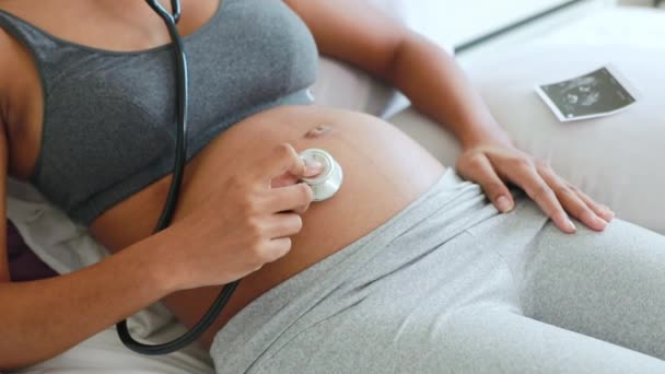怀孕的美籍泰裔母亲高兴地使用医用听诊器 倾听胎儿的心跳 并兴奋地将超声波图像放在婴儿在子宫中移动时的床上 — 图库视频影像