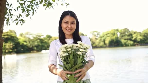 肖像画美しいかわいい穏やかな健康で陽気なアジアの女性は庭の屋外で際立っており 励ましと肯定的な願いとしてあなたに白い花を保持します — ストック動画