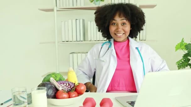 可爱的非洲裔美国女营养师坐在桌子上 微笑着举手致意 坐在医院办公室的桌子上 — 图库视频影像