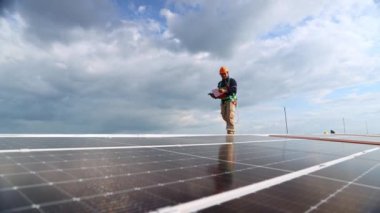 Metal çatılı endüstriyel fabrikanın güneş panellerini inceleyen Asyalı erkek elektrik mühendisi elinde matkap tutarak etrafta dolaşıyor. Panodaki her güneş panelinin önemli noktalarını kontrol ediyor..
