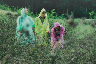 Yağmur mevsiminde seyahat eden ve yağmurluk giyen beyaz erkek ve kadınlar, Tayland tropikal ormanlarında yürüyüş çubukları kullanarak güzel şelaleleri aramak için doğa patikalarını kullanırlar..