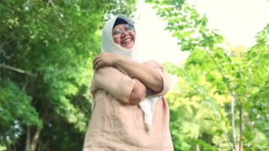 Yaşlı Asyalı Müslüman kadın tesettüre girmiş olumlu duygular besliyor serin esintiye sarılıyor güzel esinti yeşil ağaçlar gölgeli ağaçlar güzel hava zihinsel sağlığa iyi geliyor..