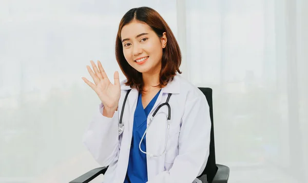 Female doctor volunteer surgeon white uniform with gentle medical stethoscope sits waving hello in a chair in the office relaxing smiling at the camera friendly to everyone.