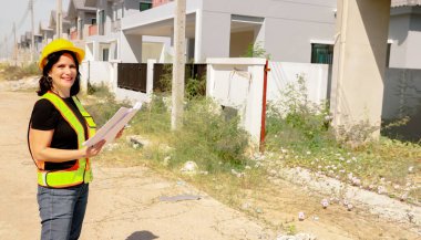 Üst düzey kadın mimar proje tasarısını elinde tutuyor. Projenin önündeki inşaat sahasında duruyor. İnşaatın ilerlemesine bakıyor..