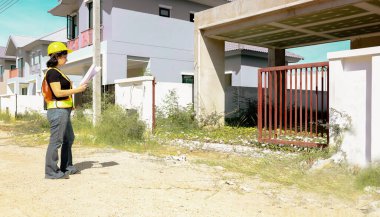 Üst düzey kadın mimar proje tasarısını elinde tutuyor. Projenin önündeki inşaat sahasında duruyor. İnşaatın ilerlemesine bakıyor..