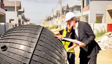 Erkek mimar ve kadın ustabaşı evin içindeki foseptiği ve kiri inceliyor ve inşaat halindeki yerli atık su arıtma projesinin planına bakıyor..