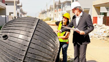 Erkek mimar ve kadın ustabaşı evin içindeki foseptiği ve kiri inceliyor ve inşaat halindeki yerli atık su arıtma projesinin planına bakıyor..