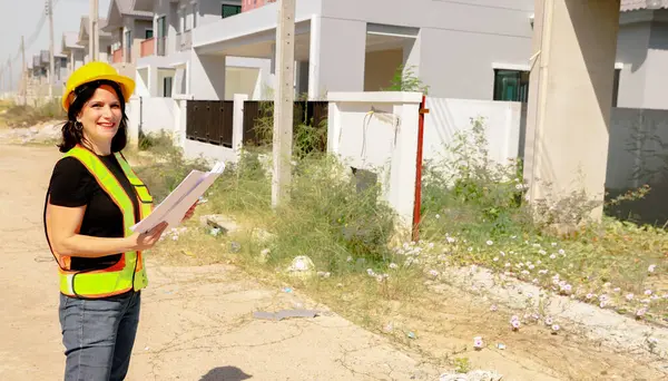 Üst düzey kadın mimar proje tasarısını elinde tutuyor. Projenin önündeki inşaat sahasında duruyor. İnşaatın ilerlemesine bakıyor..