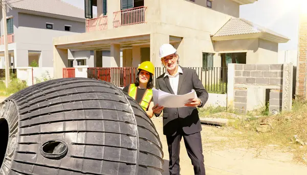 Erkek mimar ve kadın ustabaşı evin içindeki foseptiği ve kiri inceliyor ve inşaat halindeki yerli atık su arıtma projesinin planına bakıyor..