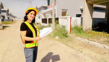 Üst düzey kadın mimar proje tasarısını elinde tutuyor. Projenin önündeki inşaat sahasında duruyor. İnşaatın ilerlemesine bakıyor..