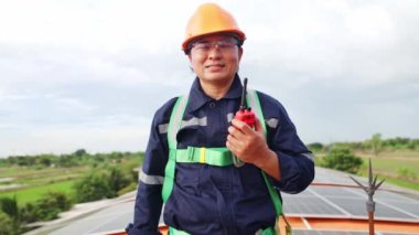 Asyalı elektrik mühendisinin portresi, endüstriyel bir fabrikanın çelik çatısında güneş panelleri kurulumunu denetliyor, bir telsiz taşıyor, nitelikli bir ekiple sahada çalışıyor..