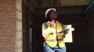 Siyahi kadın mimar, güvenlik kaskı takmış, Afro-Amerikan gülümseyen, elinde dijital tablet ve planlarla inşaat alanındaki denetim çalışmalarını kontrol ediyor..