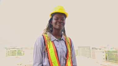 Siyahi Afro-Amerikan kadın işçilerin portresi inşaat alanında çalışıyor uzun saç örgüleri, başlık takıyor benzersiz bir cazibeyle gülümsüyor inşaat alanında iyi ruh hali içinde iş arkadaşlarını selamlıyor..