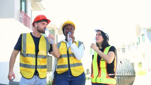 建設現場のさまざまな国籍の男性と女性の労働者のグループが立ち 休憩中にさわやかな笑顔で喉を癒すためにボトルにきれいな蒸留水を飲んだ — ストック動画