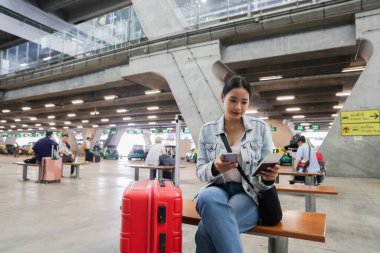 Elinde bavuluyla oturan Asyalı kadın, uluslararası havaalanındaki bir terminalde pasaport ve uçak biletleri gibi belgelerin geçerliliğini kontrol ediyor..