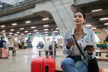 Elinde bavuluyla oturan Asyalı kadın, uluslararası havaalanındaki bir terminalde pasaport ve uçak biletleri gibi belgelerin geçerliliğini kontrol ediyor..