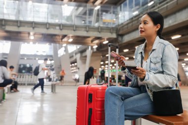 Elinde bavuluyla oturan Asyalı kadın, uluslararası havaalanındaki bir terminalde pasaport ve uçak biletleri gibi belgelerin geçerliliğini kontrol ediyor..
