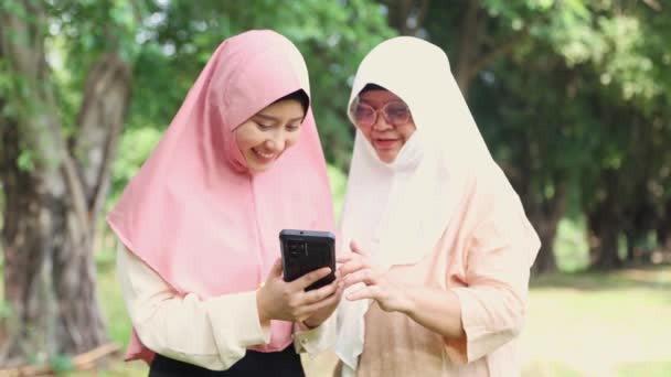 陽気な家族2人のイスラム教徒の母親と娘は日陰の庭を歩き 娘たちがスマートフォンのカメラから彼女の写真を見せたことを喜び 良い気分が家族関係を強化しました — ストック動画