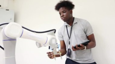 Genç siyahi erkek öğrenci, dijital tablete robot verileri kaydediyor. Sınıf laboratuvarında, etkin sistem üzerinde araştırma ve geliştirme yapan endüstriyel robot kol mühendisi..