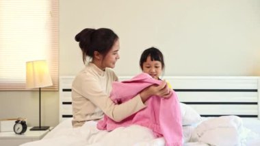 Güzel anne ve Asyalı kız yatak odasındaki yatakta oturup parlak pembe bir kazak hazırlıyorlar ve küçük kızları yarın anaokulunda giymeye çalışıyor..