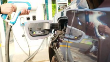 EV elektrikli araba kullanan genç bir kadın, benzin istasyonundaki gri ve siyah elektrikli akü prizine takılıyor: Temiz enerji aracı Alternatif enerji popüler bir kavram haline geliyor.